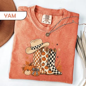 May include: A yam-colored t-shirt featuring a graphic print of a cowboy hat, three western-style boots, and pumpkins. The boots and hat display checkered patterns. The shirt has a crew neckline and a Comfort Colors brand label at the collar.