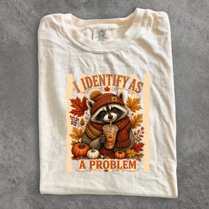 May include: A cream-colored t-shirt featuring a graphic of a raccoon wearing a brown knit hat and scarf, holding a pumpkin-themed drink. The text reads I IDENTIFY AS A PROBLEM, surrounded by autumn leaves, berries, and pumpkins in orange and brown.