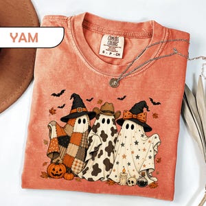 May include: A coral-colored t-shirt featuring a graphic of three ghosts. One ghost wears a witch hat and a patchwork blanket, one wears a cowboy hat and cow print, and one wears a witch hat with a bow and star pattern. Includes pumpkins, bats, and leaves.
