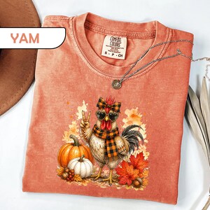 May include: A coral-colored t-shirt featuring a graphic of a chicken wearing a plaid scarf, a matching bow, and heart-shaped sunglasses. The chicken is making a peace sign next to pumpkins, autumn leaves, and acorns. The shirt has a crew neck.