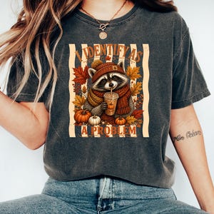 May include: A charcoal gray t-shirt featuring a graphic of a raccoon wearing a knit hat and scarf, holding a pumpkin spice drink. The design includes autumn leaves, pumpkins, and the text "I IDENTIFY AS A PROBLEM" in a bold, orange font.