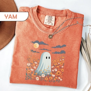 May include: A yam-colored short-sleeve t-shirt featuring a graphic of a white ghost standing in a field of yellow and white flowers under a moon and dark clouds. The shirt has a Comfort Colors brand label at the neck and a crew neckline.