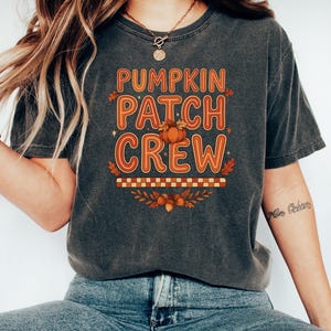 May include: A charcoal grey short-sleeve t-shirt featuring the orange text PUMPKIN PATCH CREW, a small pumpkin illustration, a checkered pattern, and autumn leaves and acorns printed on the front center of the garment.