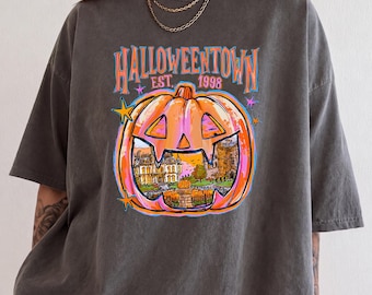 May include: A charcoal grey t-shirt featuring a graphic of a large orange jack-o'-lantern with a detailed illustration of a house inside. The text reads HALLOWEENTOWN EST. 1998 in orange and blue, accented by small pink and yellow stars.