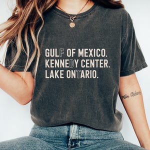 May include: A charcoal grey short-sleeve t-shirt featuring the white text GULF OF MEXICO. KENNEDY CENTER. LAKE ONTARIO. The shirt has a crew neckline and a washed, vintage-style finish. The letters F, D, and T are outlined in a thin white border.