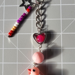 Könnte beinhalten: Ein silberner Schlüsselanhänger mit einem Sternanhänger, einem farbigen Perlenstab, einem herzförmigen Anhänger mit einer rosa Perle, einer rosa Kugel und einem kleinen rosa Entenanhänger. Der Schlüsselanhänger liegt auf einer grauen Rasteroberfläche.
