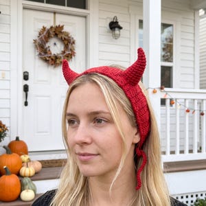 May include: A bright red crocheted headband featuring two prominent, upright devil horns. The accessory is constructed with a textured yarn stitch pattern and includes long, thin ties at the bottom to secure the piece around the head.