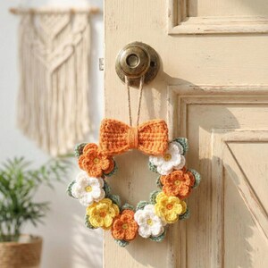 May include: A handmade crochet wreath with a bow and flower design. The wreath features orange, yellow, and white flowers with green leaves. The bow is a vibrant orange color. The wreath is hanging on a cream-colored door.