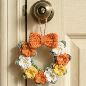 May include: A handmade crochet wreath with orange, yellow, and white flowers, and green leaves. The wreath is adorned with an orange bow and is suspended from a door knob. The wreath is a charming, decorative piece.