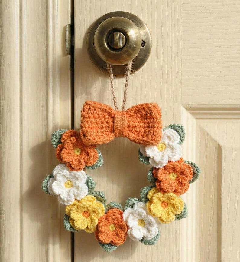 May include: A handmade crochet wreath with orange, yellow, and white flowers, and green leaves. The wreath is adorned with an orange bow and is suspended from a door knob. The wreath is a charming, decorative piece.
