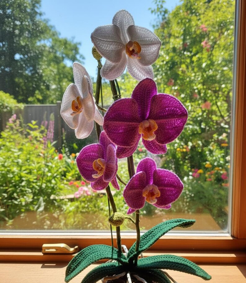 May include: A handcrafted crochet orchid plant with white and purple flowers. The flowers have yellow centers and are arranged on green stems with green leaves. The plant sits in front of a window with a blurred outdoor background.