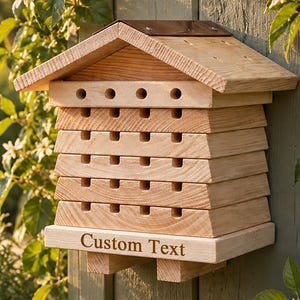 Può includere: Una casetta per api in legno con tetto a due spioventi e molteplici camere di nidificazione orizzontali dotate di fori d'ingresso circolari. La struttura presenta una base piatta con la scritta Custom Text incisa sulla parte anteriore.