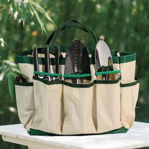 May include: A beige and green garden tool tote bag with multiple pockets and a handle. The bag contains various gardening tools, including a trowel, hand rake, and a weeding knife. The background features lush green bamboo.