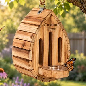 May include: A teardrop-shaped wooden butterfly house with a natural wood grain finish, featuring three vertical entry slots, a small circular feeding tray, and the text Custom Text engraved on the front, suspended by a thick rope from a tree branch.