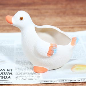 May include: A white ceramic duck planter with orange beak and feet, and blue and orange accents on the wings. The planter is sitting on a newspaper.