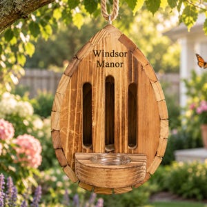 May include: A teardrop-shaped wooden insect hotel with a dark stained finish, featuring three vertical entry slots and a small circular glass dish at the base. The text "Windsor Manor" is engraved on the upper front section of the wooden structure.