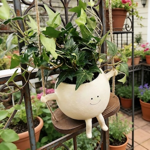 Peut inclure: Un pot de plante en céramique blanc cassé fantaisiste en forme de personnage, avec un visage souriant, des bras et des jambes, suspendu à une balançoire en bois marron. Le pot est rempli de lierre vert luxuriant. Le fond montre un jardin.