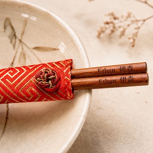 Könnte beinhalten: Ein Paar braune Holzstäbchen, graviert mit dem Namen Ethan und den japanischen Schriftzeichen 伊森, teilweise in einer roten Stoffhülle mit goldenem geometrischem Muster und einem dekorativen roten Knotenknopf.