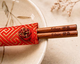 Könnte beinhalten: Ein Paar braune Holzstäbchen, graviert mit dem Namen Ethan und den japanischen Schriftzeichen 伊森, teilweise in einer roten Stoffhülle mit goldenem geometrischem Muster und einem dekorativen roten Knotenknopf.