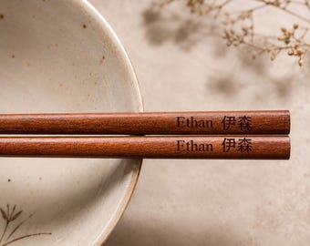 Könnte beinhalten: Ein Paar glatte, dunkelbraune Essstäbchen aus Holz mit der eingravierten Aufschrift Ethan 伊森 auf jedem Stäbchen. Die Stäbchen liegen horizontal auf dem Rand einer hellen Keramikschale mit einer dezent gesprenkelten Textur.