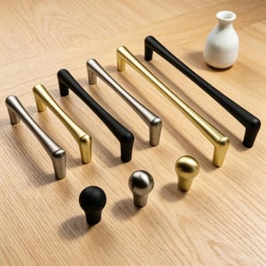 May include: A collection of modern cabinet hardware, including silver, gold, and black handles and knobs. The handles are bar-shaped, and the knobs are round. A small white vase is also present.