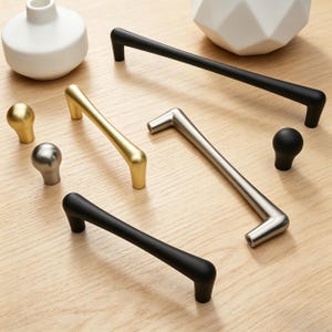 May include: A collection of cabinet hardware, including black, gold, and silver handles and knobs. The handles are bar-shaped and the knobs are spherical. The hardware is displayed on a light wood surface.