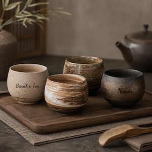 May include: Four ceramic tea cups with distinct finishes arranged on a wooden tray. One cup is light beige with the text Sarah's Tea, one has brown and white swirls, one is textured brown, and one is dark charcoal with the text Neuer.