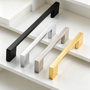 May include: Four cabinet pulls in black, silver, brushed nickel, and gold. Each pull has a rectangular bar and square ends. The pulls are displayed on a white shelf.