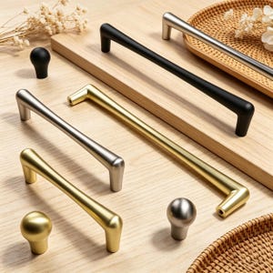 May include: A collection of cabinet hardware, including gold, silver, and black handles and knobs. The handles are bar-style, and the knobs are spherical. The hardware is displayed on a light wood surface, with a woven tray in the background.