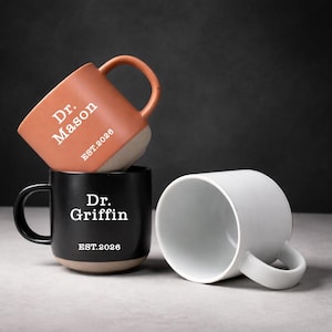 Könnte beinhalten: Drei Keramikbecher gestapelt. Der obere Becher ist orange mit dem Text "Dr. Mason EST.2026". Der mittlere Becher ist schwarz mit dem Text "Dr. Griffin EST.2026". Der untere Becher ist weiß.