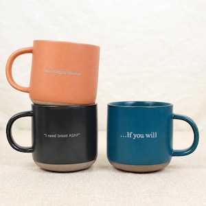 Può includere: Tre tazze in ceramica arancione, nera e verde acqua. Ogni tazza ha una base arrotondata e un manico grande. La tazza arancione ha il testo "the charged tension". La tazza nera dice "I need bread ASAP". La tazza verde acqua dice "...If you will".