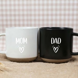 May include: Two ceramic mugs, one white and one black, with brown bases. The white mug has the word "MOM" and a heart, while the black mug says "DAD" with a heart. Both mugs have a simple, modern design.