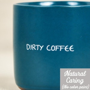 Könnte beinhalten: Eine türkisfarbene Keramiktasse mit den Worten "DIRTY COFFEE" in weißer Schrift. Die Tasse hat eine abgerundete Form und einen braunen Boden. Die Worte "Natural Caring (No color paint)" sind ebenfalls sichtbar.