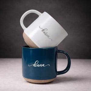 Puede incluir: Dos tazas de cerámica apiladas una encima de la otra. La taza inferior es azul oscuro con el nombre "Diana" en escritura blanca. La taza superior es blanca con el nombre "Karin" en escritura blanca. Ambas tazas tienen una base beige.