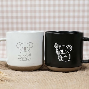 May include: Two ceramic mugs, one white and one black, each featuring a koala bear design. The white mug has a seated koala, while the black mug shows a koala hugging a branch. Both mugs have a brown base.