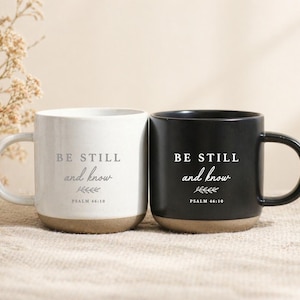 Puede incluir: Dos tazas de cerámica, una blanca y otra negra, cada una con el texto BE STILL and know, un pequeño gráfico de hoja y la cita PSALM 46:10. Ambas tazas tienen un acabado superior liso y una base de color tostado sin esmaltar con asa.