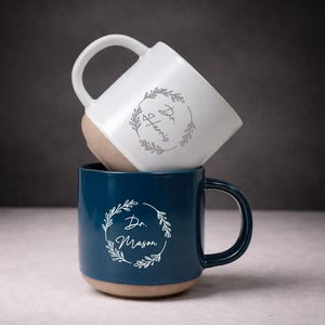 Puede incluir: Dos tazas de cerámica, una blanca y otra azul marino, apiladas. Cada taza presenta un diseño de corona botánica con el texto "Dr. Harris" y "Dr. Mason". La base de cada taza es de color beige natural.