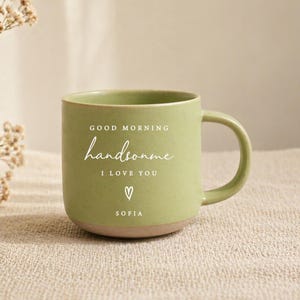 Puede incluir: Una taza de cerámica de color verde claro con un acabado moteado y un asa en el lado derecho. El frente presenta texto blanco que dice GOOD MORNING handsome I LOVE YOU, seguido de un pequeño icono de corazón y el nombre SOFIA impreso abajo.