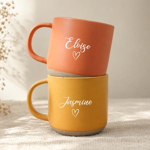 May include: Two stacked ceramic mugs. The top mug is orange with the name "Eloise" and a heart. The bottom mug is yellow with the name "Jasmine" and a heart. Both mugs have a grey base and a curved handle.
