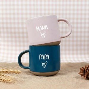 May include: Two ceramic mugs stacked on top of each other. The top mug is light pink with the word "MAMA" and a heart symbol in white. The bottom mug is dark blue with the word "PAPA" and a heart symbol in white.