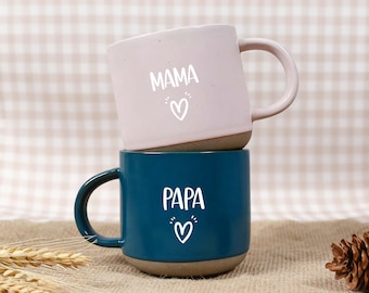 May include: Two ceramic mugs stacked on top of each other. The top mug is light pink with the word "MAMA" and a heart symbol in white. The bottom mug is dark blue with the word "PAPA" and a heart symbol in white.