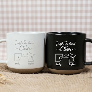 Pode incluir: Duas canecas de cerâmica, uma branca e outra preta, com bases castanhas. Cada caneca tem o texto "I wish we lived Closer" e um gráfico de mapa com nomes. A caneca branca tem uma alça branca, e a caneca preta tem uma alça preta.