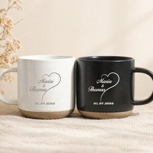 May include: Two ceramic mugs, one white and one black, with a heart design and the names "Maria & Thomas" and the date "01.07.2026" printed on them. The mugs have a beige base and a curved handle.