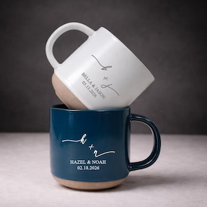 Puede incluir: Dos tazas de cerámica, una blanca y otra azul marino, apiladas. La taza blanca tiene los nombres "Bella & Jason" y la fecha "03.13.2026". La taza azul tiene los nombres "Hazel & Noah" y la fecha "02.18.2026". Ambas tazas tienen una base beige.