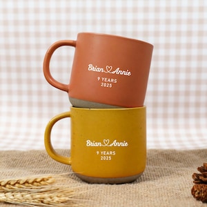 Puede incluir: Dos tazas de cerámica apiladas, una naranja y otra amarilla. La taza naranja tiene texto blanco que dice "Brian & Annie 9 YEARS 2025". La taza amarilla tiene el mismo texto. Ambas tazas tienen un asa redonda.