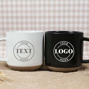 May include: Two ceramic mugs, one white and one black, with brown bases and large handles. The white mug has the text "YOUR TEXT HERE" in a circle, and the black mug has "YOUR LOGO HERE". Both mugs are on a burlap surface.