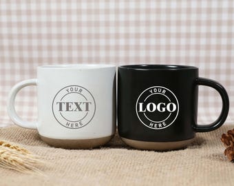 May include: Two ceramic mugs, one white and one black, with brown bases and large handles. The white mug has the text "YOUR TEXT HERE" in a circle, and the black mug has "YOUR LOGO HERE". Both mugs are on a burlap surface.