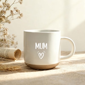 May include: White ceramic mug with a brown base, featuring the word "MUM" in white script, along with a heart symbol. The mug has a curved handle and is set against a neutral background with dried flowers.