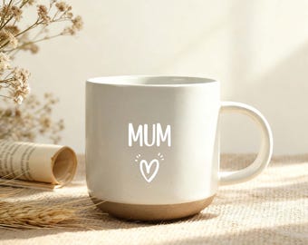 May include: White ceramic mug with a brown base, featuring the word "MUM" in white script, along with a heart symbol. The mug has a curved handle and is set against a neutral background with dried flowers.