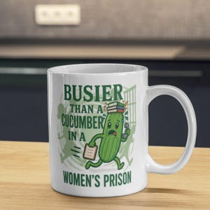 Könnte beinhalten: Weiße Keramik-Tasse mit humorvoller Grafik. Das Design zeigt eine Cartoon-Gurke, die mit Büchern, einem Telefon und einem Notizblock rennt, mit dem Text "BUSIER THAN A CUCUMBER IN A WOMEN'S PRISON". Die Tasse hat einen gebogenen Griff.